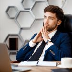 Young successful businessman sitting at workplace, office background.